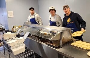 Three men in a room with blue walls serving food onto cafeteria trays