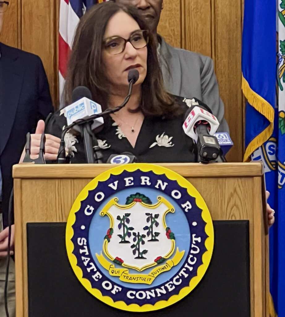 CEO Kara Capone speaking at the Governor's podium, with the seal of the State of Connecticut on the front above a black box.